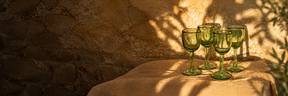 Copas verdes de vidrio soplado de The Blown-Glass Collection sobre lino y muro de piedra con luz natural.