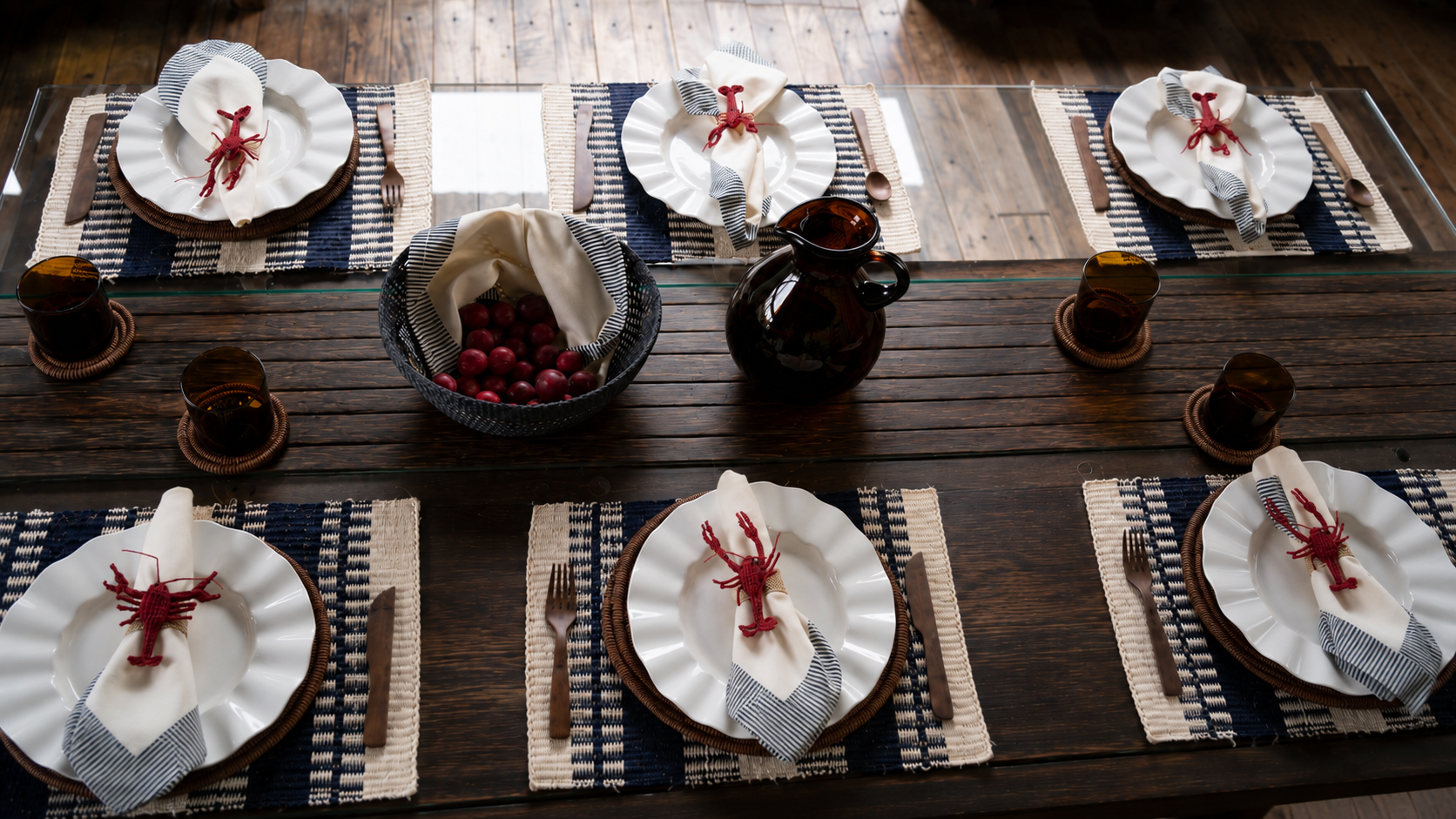 Mesa puesta con individuales tejidos, platos blancos, servilleteros de langosta en palma de iraca y cristalería ámbar.