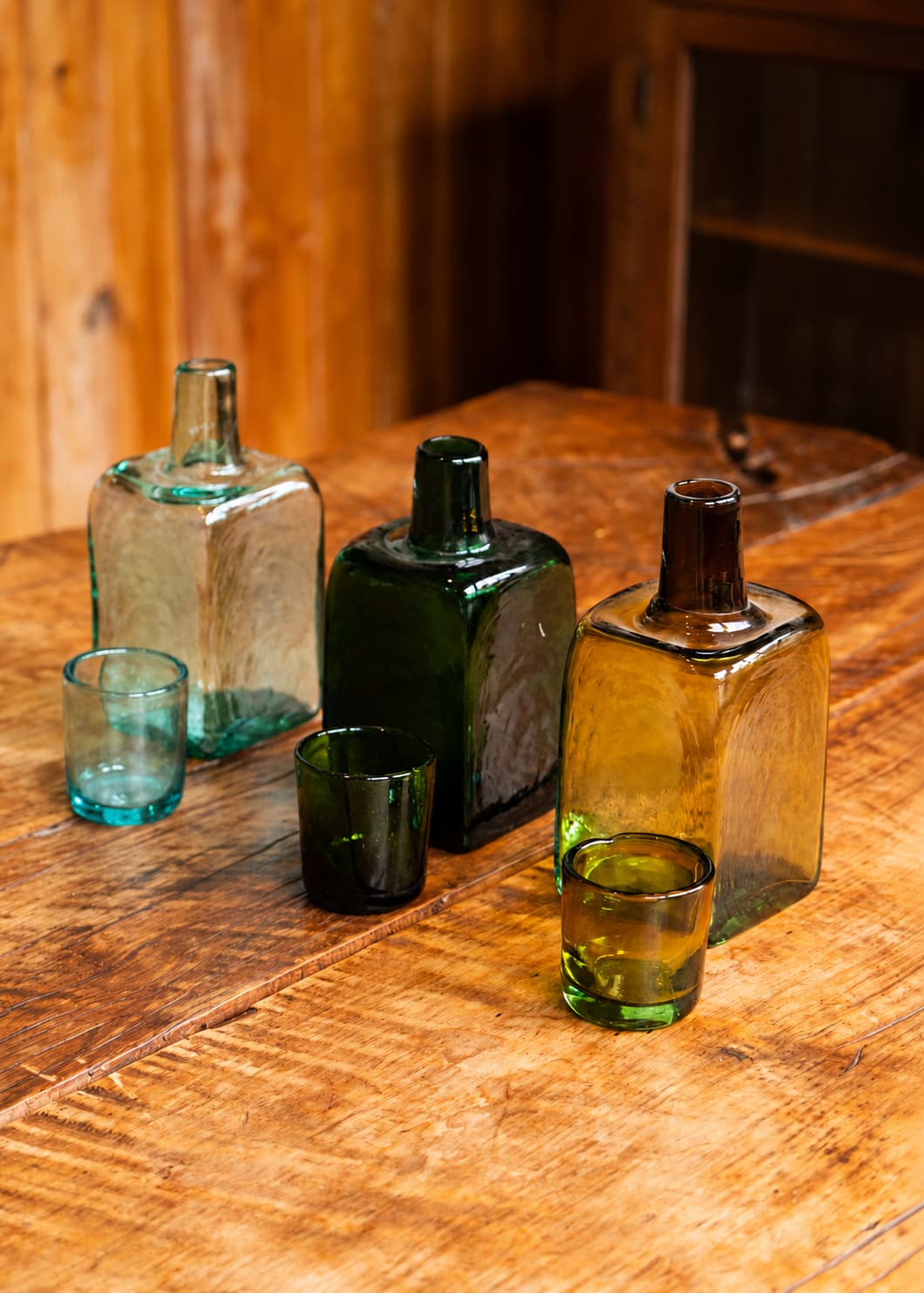 Tres botellas para nochero de vidrio soplado en azul agua, verde y ámbar, cada una con su vaso, sobre una mesa de madera.