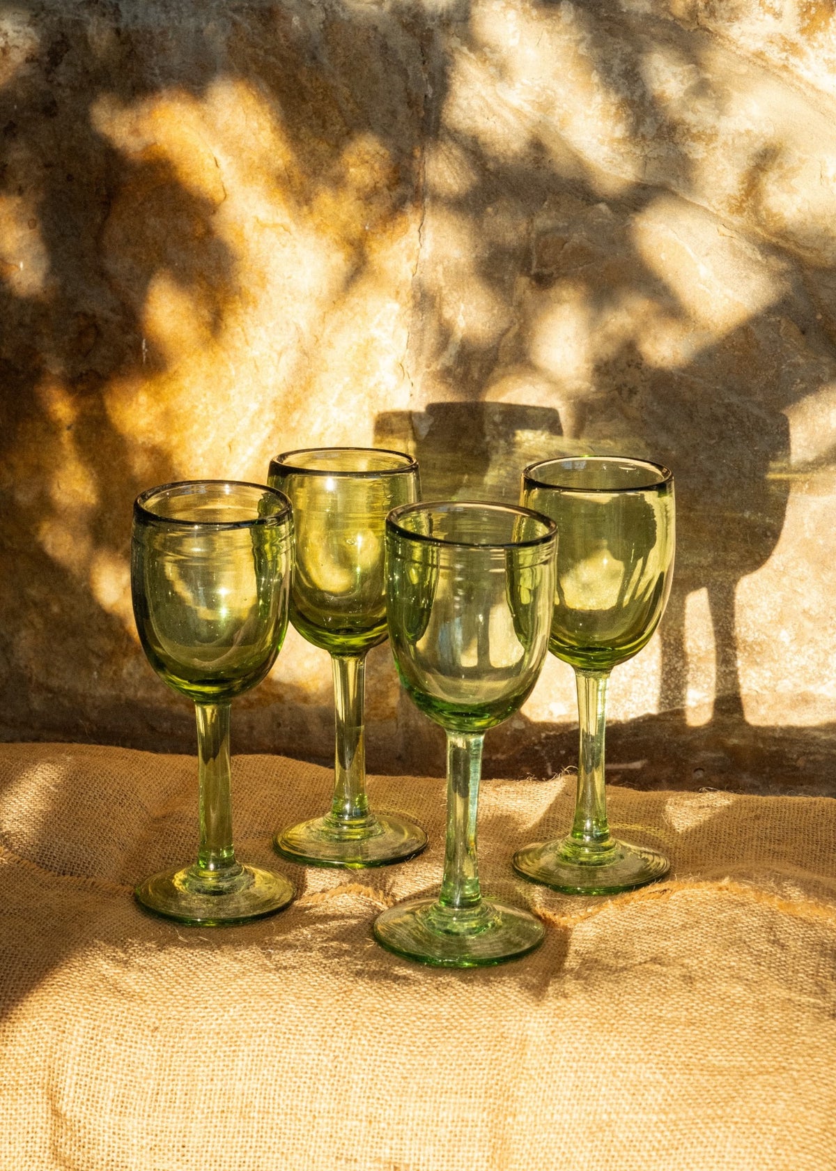 Copas de vino altas verdes de vidrio soplado sobre textil natural con luz cálida y fondo de piedra.