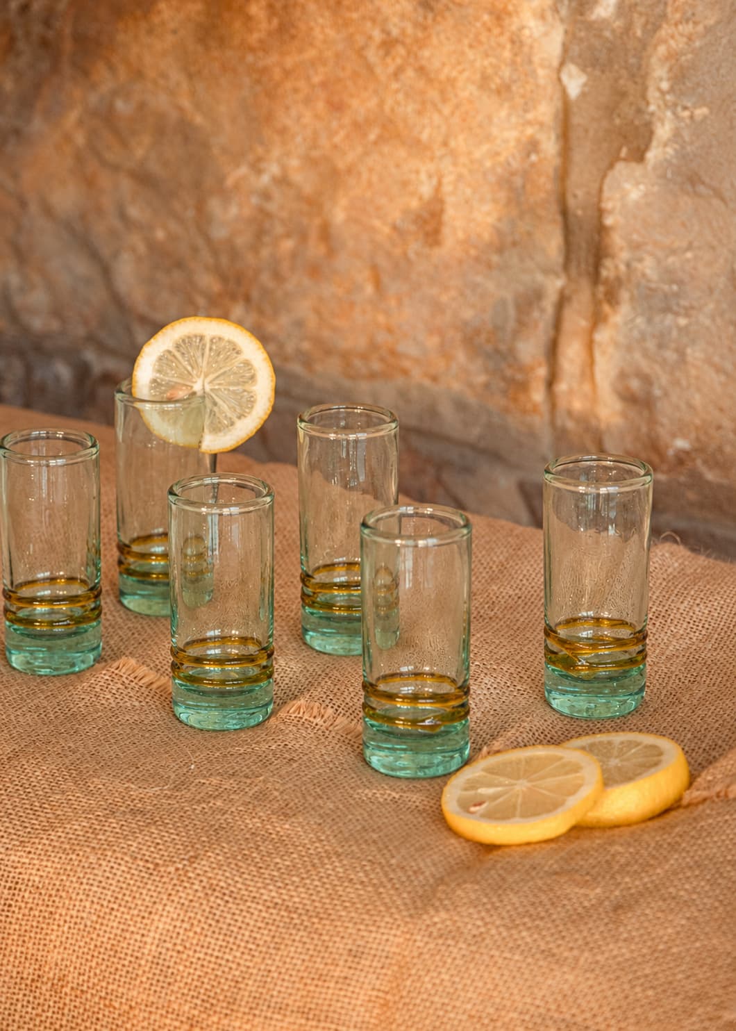 Set de seis vasos de shot con dos hilos ámbar sobre una mesa de yute con limones y muro de piedra.