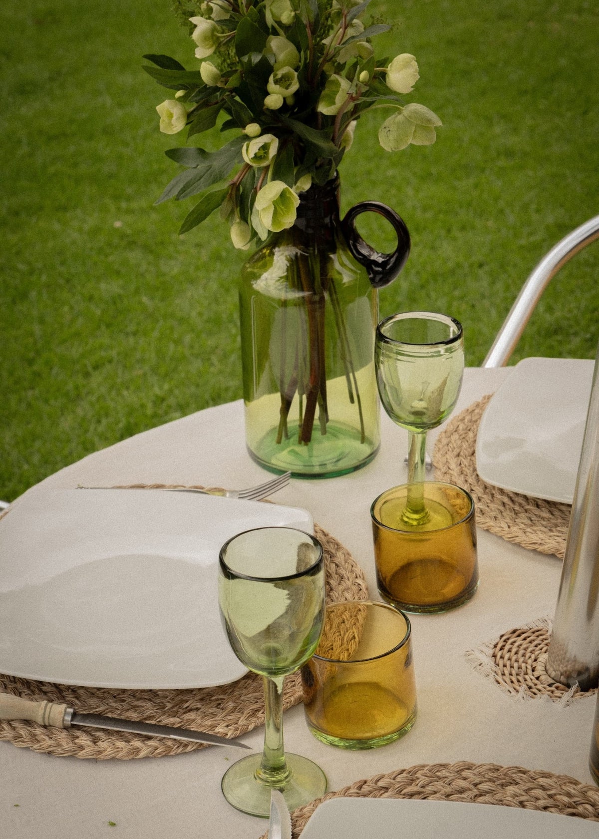 Copas de vino altas verdes de vidrio soplado en una mesa exterior con vajilla y cristalería artesanal.