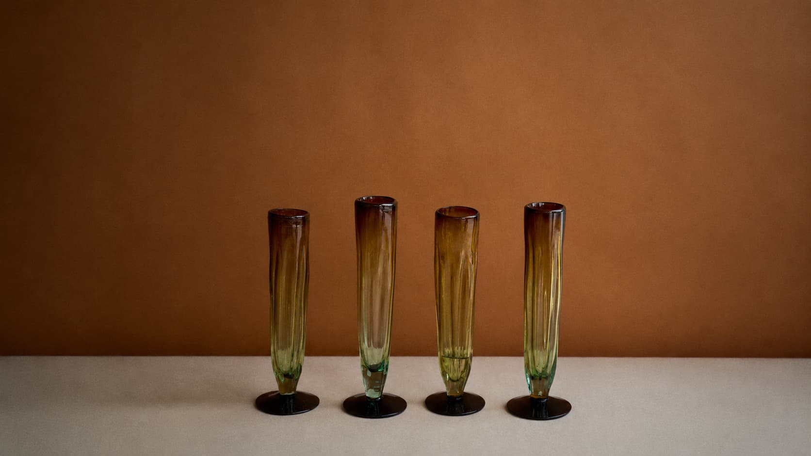 Copas de vidrio reciclado ámbar y verde dispuestas en línea sobre fondo cálido.