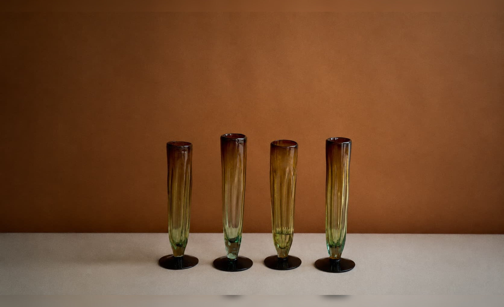 Copas de vidrio reciclado ámbar y verde dispuestas en línea sobre fondo cálido.