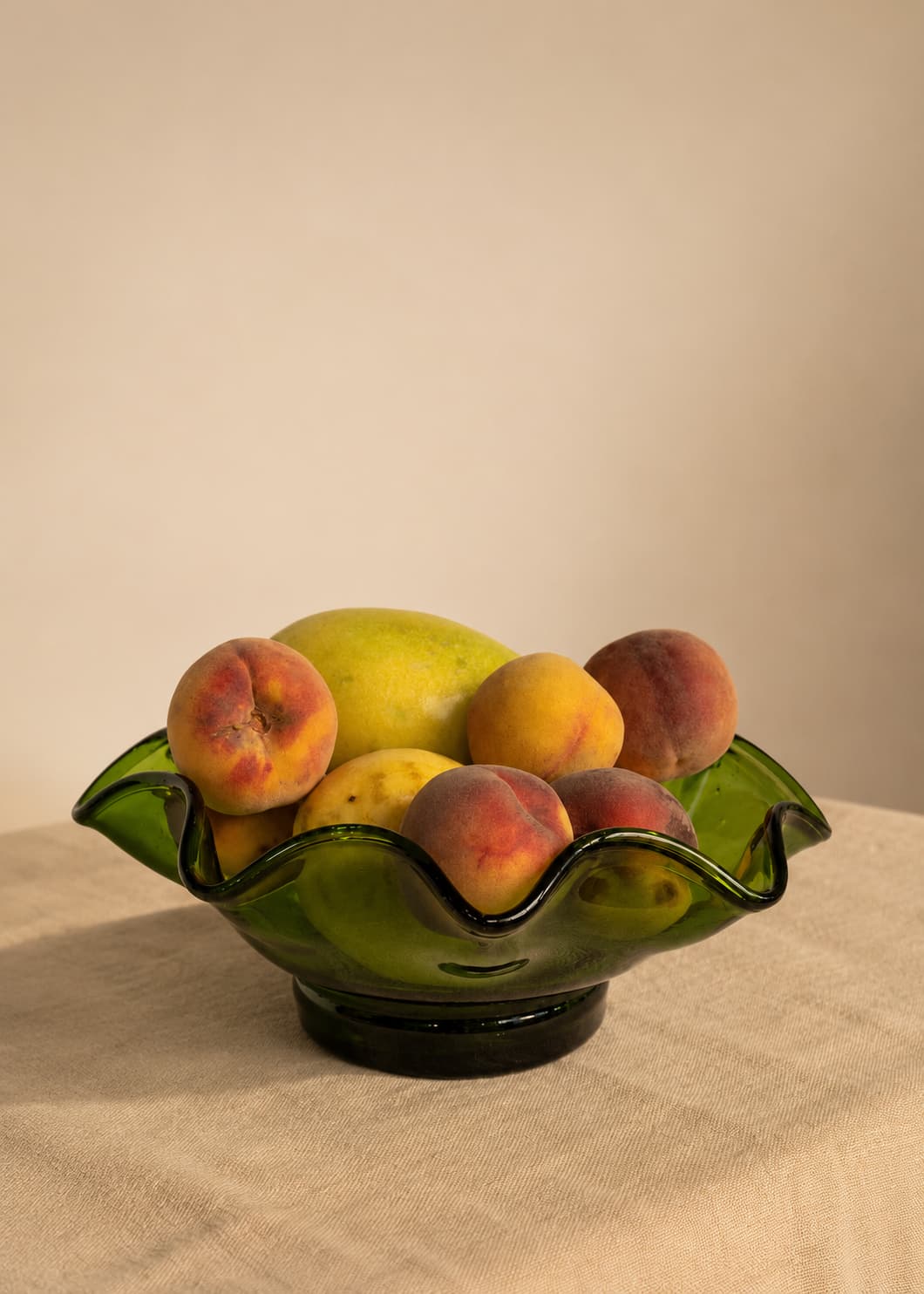 Frutero Ondulado Verde en vidrio soplado con frutas sobre superficie textil beige.