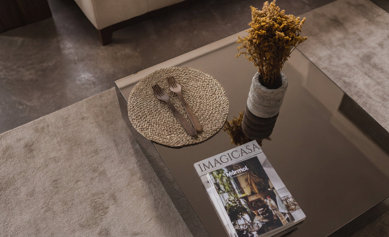 Mesa de vidrio con individual tejido en fibras naturales, cubiertos de madera, revista y arreglo floral en ambiente editorial.