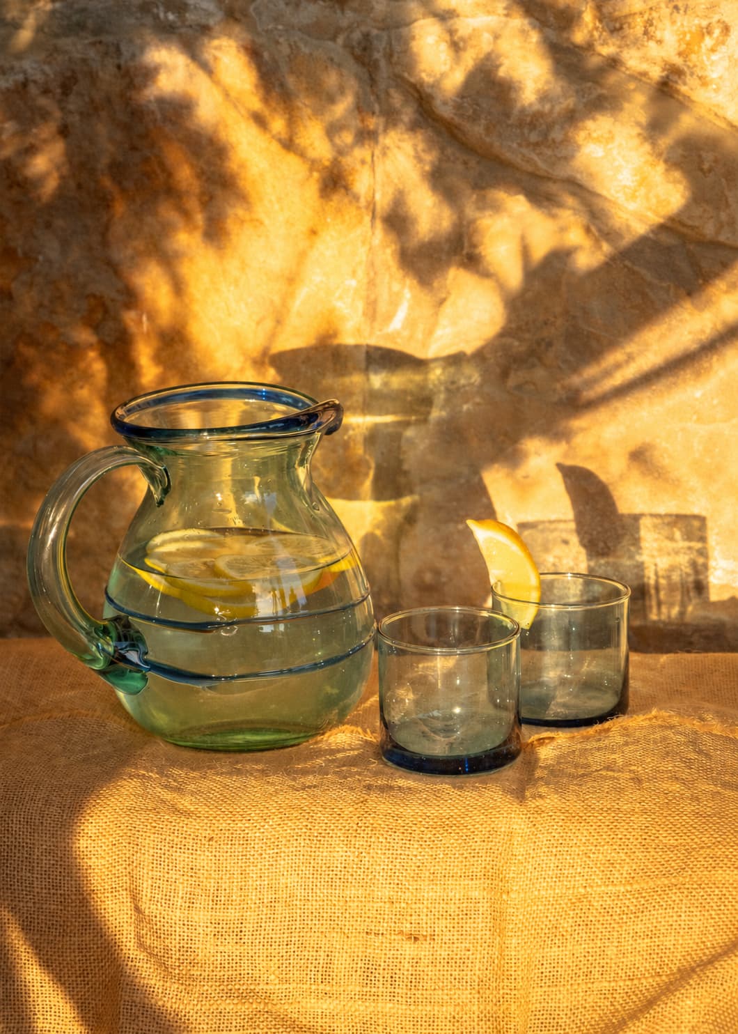 Jarra Dos Hilos en vidrio soplado con vasos, iluminada por luz natural.