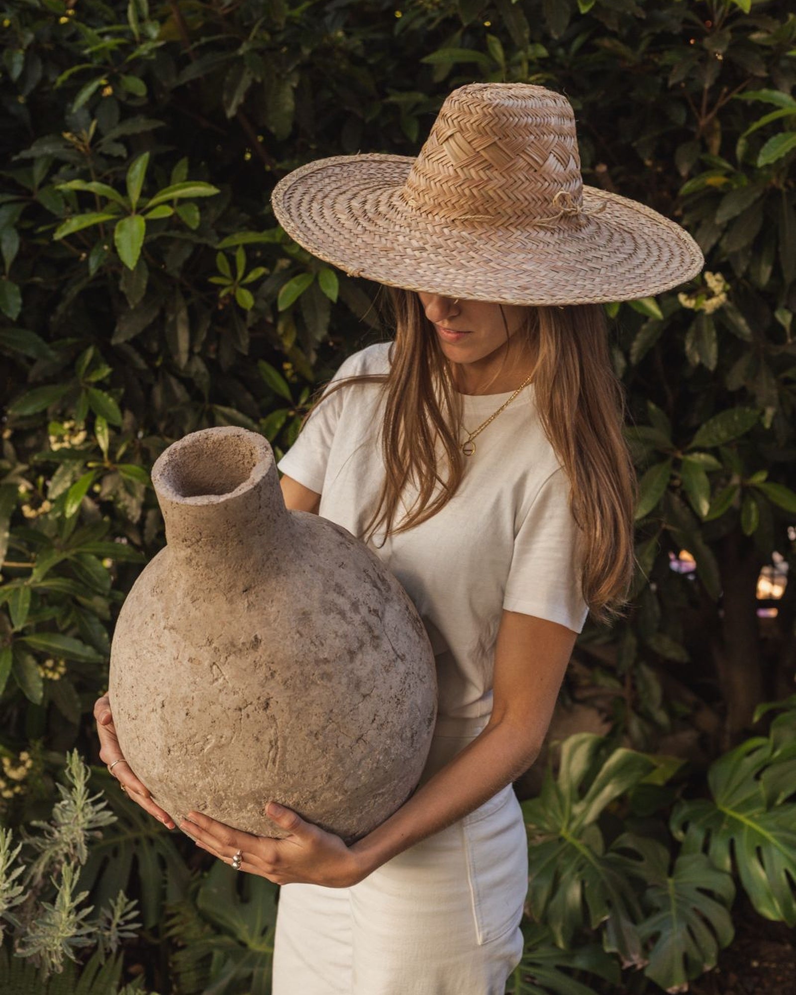Mujer con sombrero de palma sosteniendo una vasija artesanal entre plantas tropicales.