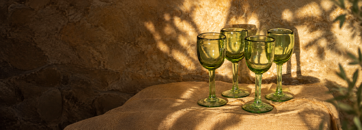 Copas verdes de vidrio soplado de The Blown-Glass Collection sobre lino y muro de piedra con luz natural.