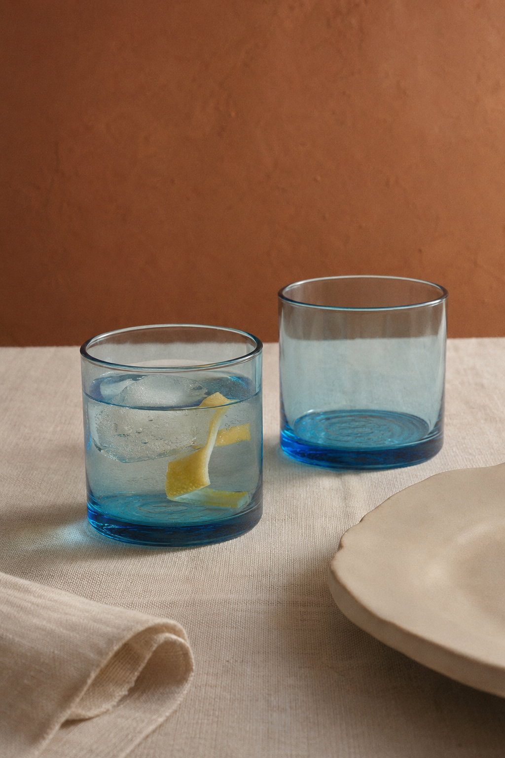 Dos vasos azules de vidrio soplado  sobre una mesa de lino; uno servido con agua fría y hielo.