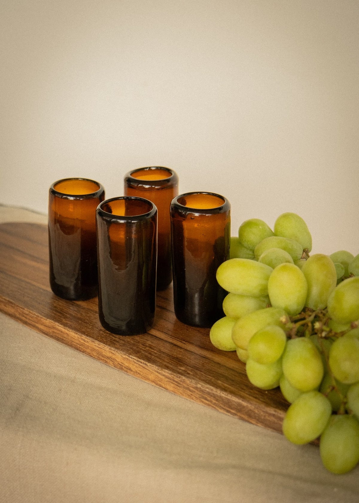 Vasos de shot artesanales en vidrio ámbar sobre tabla de madera junto a uvas verdes.